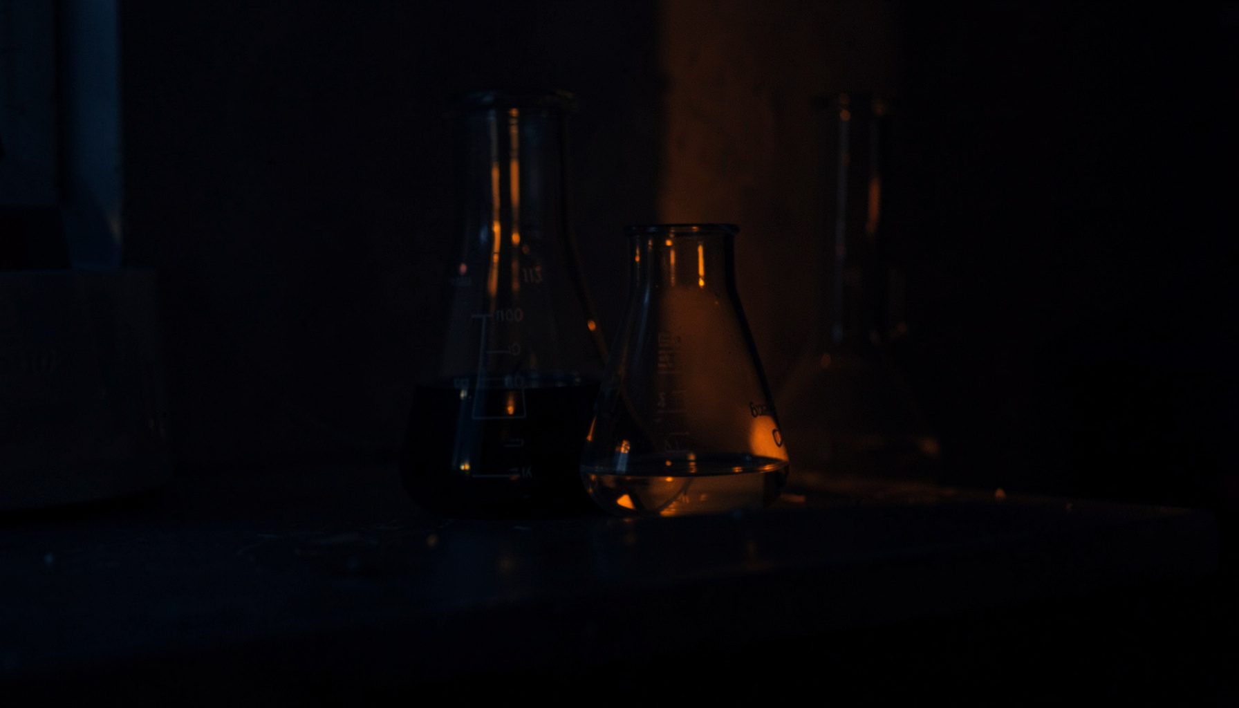 Dark laboratory glassware on a shadowy bench, deep navy and amber tones