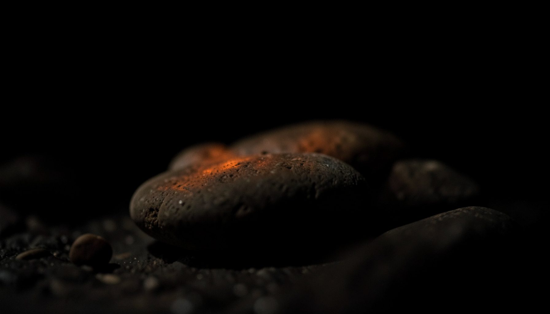 Dark, low-key photograph of smooth rounded stones in muted brown and gray tones, resting on a shadowy surface with deep black background